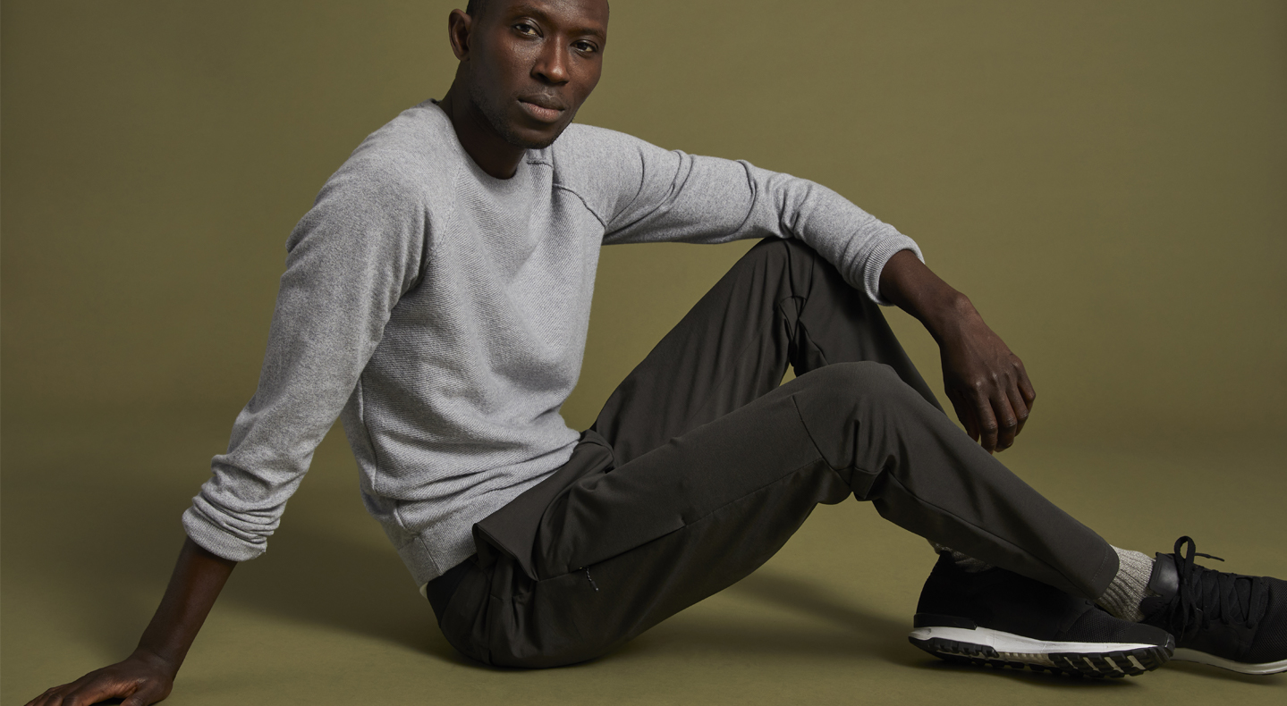 A person sits with bent knees and their hand on their knee in front of a brown/green background wearing a long sleeve white shirt from Dockers® and camo green Dockers® pants with black sneakers.