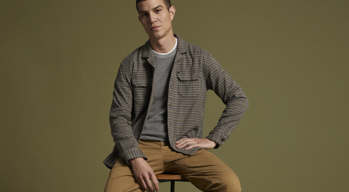 A person sits on a stool in front of a camo green background with their left hand on their left hip. They are wearin ga long sleeve grey/green button down over a t-shirt with tan khaki Dockers® pants.