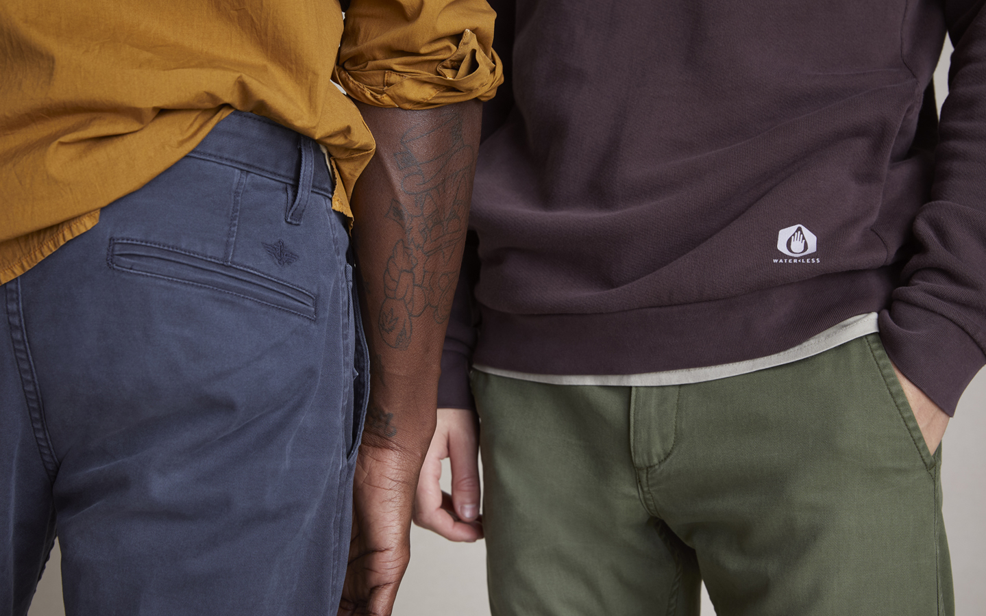 a cropped photo showing the torsos and tops of legs of two people. The person on the left faces away from the camera and wears a mustard colored top tucked into charcoal-blue pants. The person on the right faces the camera and they wear a dark brown crewneck sweatshirt and green Dockers® pants.