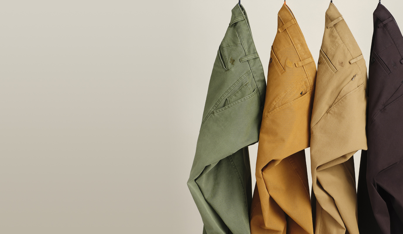 Four pairs of Dockers® pants hang by their belt loops in front of a light grey background. From left to right: green, orange, tan, black.