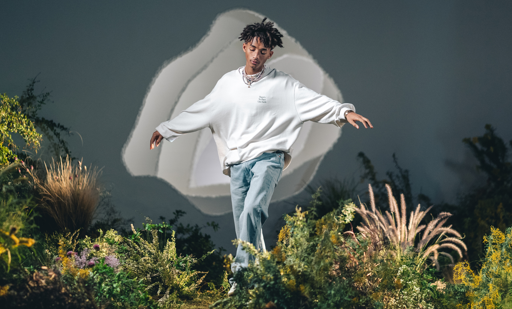 Jaden Smith poses with his eyes closed and armed stretched out wearing a stack of necklaces, a white Levi's® pullover reading "There's No Place Like Earth" and blue denim Levi's® jeans. He walks through a field of shrubbery and flowers in front of a dark sky background. An abstract amorphous white shape is behind his torso.