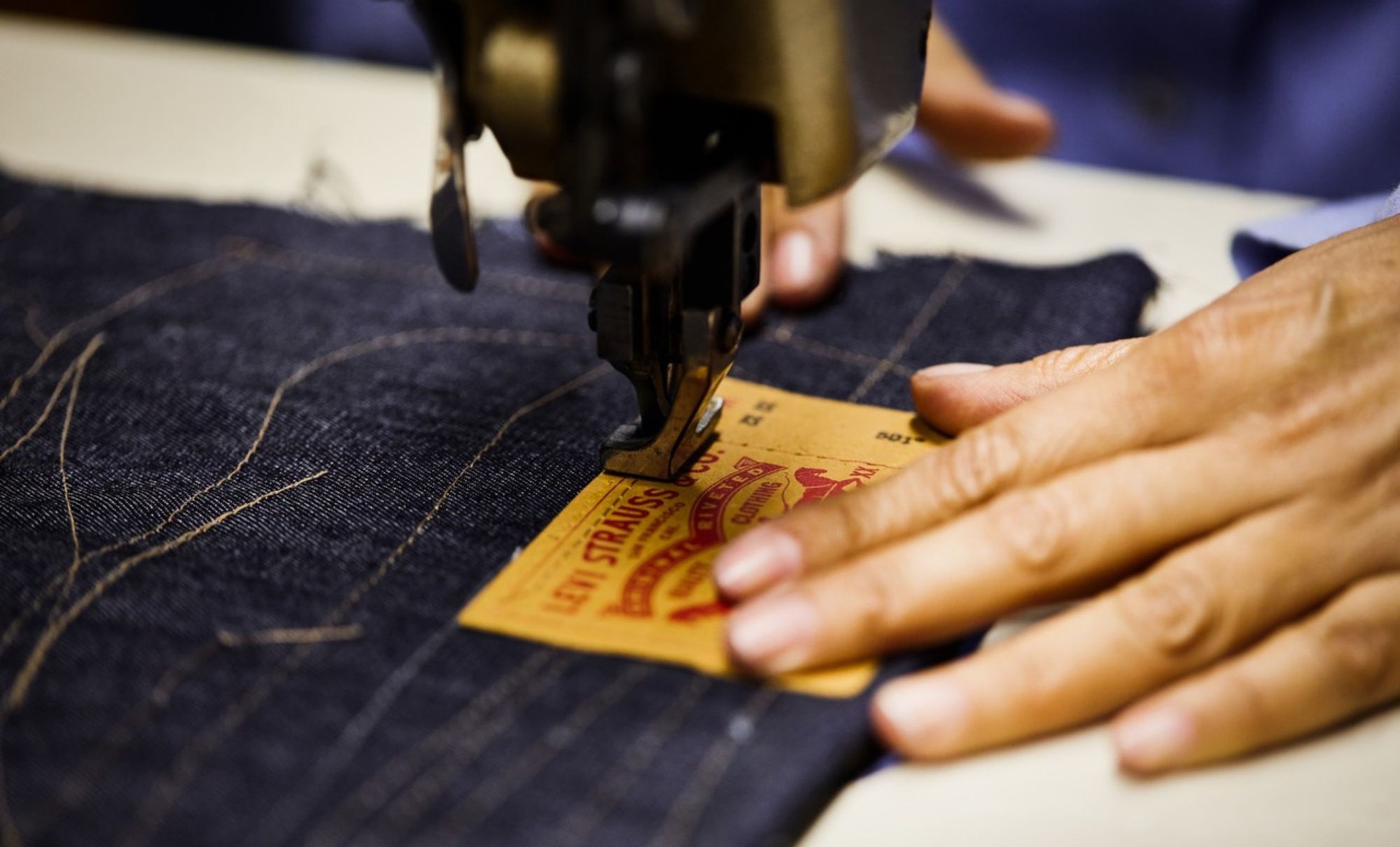 A pair of hands works at a sewing machine to sew a Levi's® leather patch onto the back of a pair of Levi's® denim jeans.