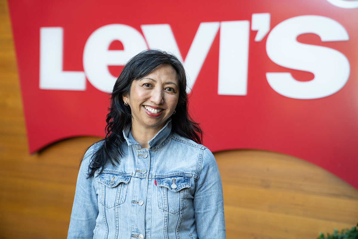 A portrait of Levi Strauss Foundation Executive Director Fatima Angeles smiling in front of a red Levi's® sign. She wears a blue Levi's® button up top.