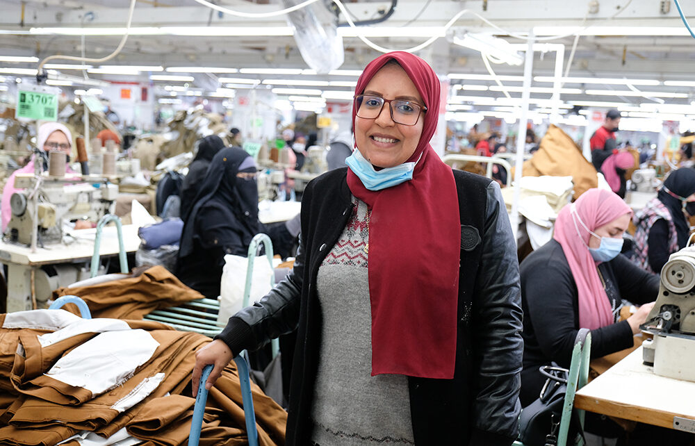 Alysayda Abdelrahim poses in a LS&Co. manufacturing facility. She wears a red scarf around her head, a black jacket and a grey top. There are people working in the background.