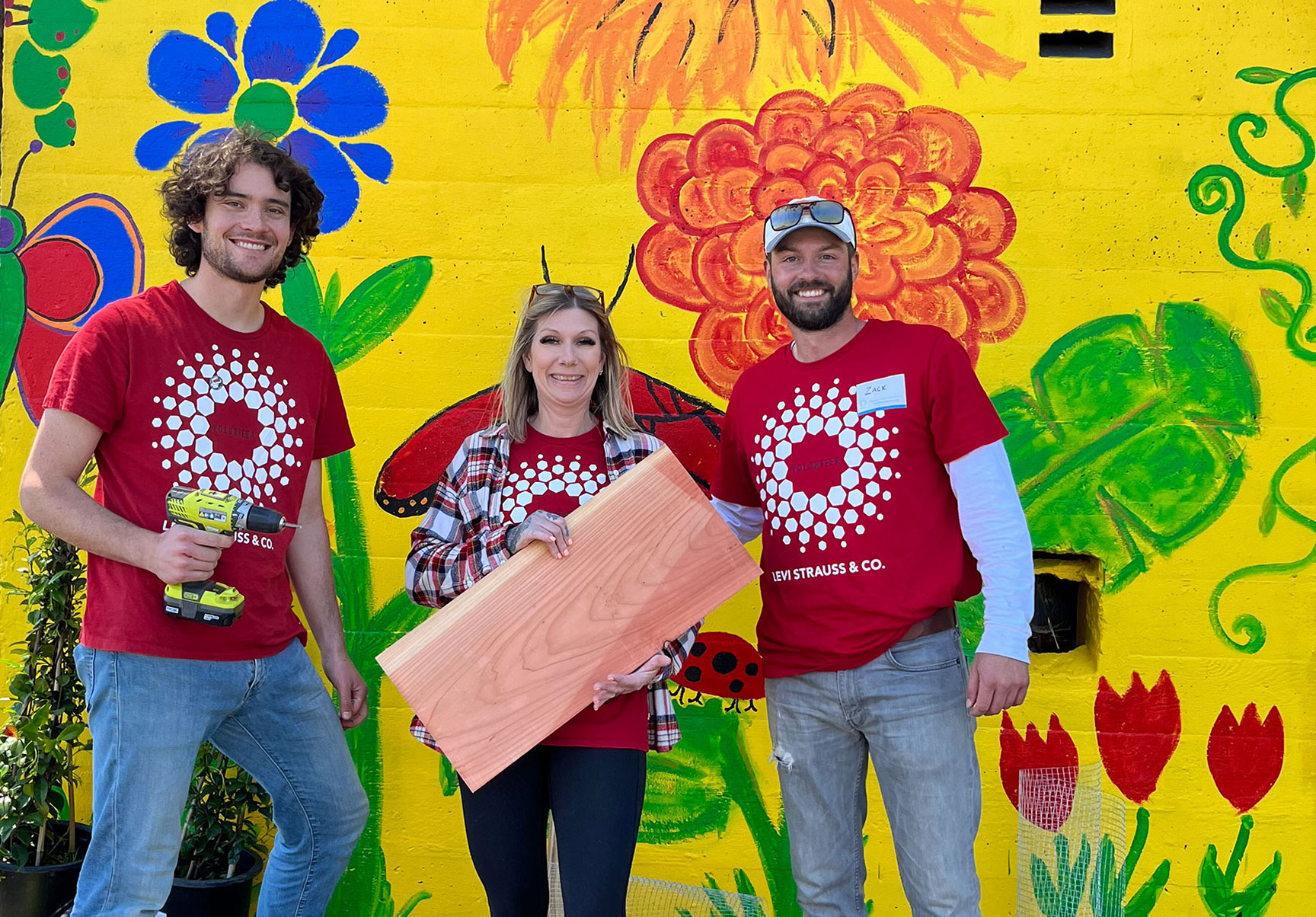 Three LS&Co. employee volunteers smile and pose in front of a yellow mural with colorful flowers painted on the wall. The employees are wearing red LS&Co. community day t-shirts. The person on the left holds a yellow electric drill and the person in the middle holds a plank of wood.