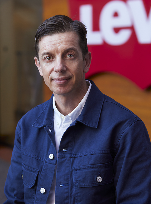 LS&Co. Chief Digital Officer Jason Gowans poses in front of a red Levi's® sign. He wears a blue button down over a white collared shirt.