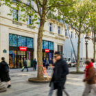 The exterior of the new Levi's® flagship store in Paris, Levi's® Champs-Élysées. A cream colored Parisian style building with large windows and the red Levi's® batwing logo placed throughout. Trees line the sidewalk and people walk around.