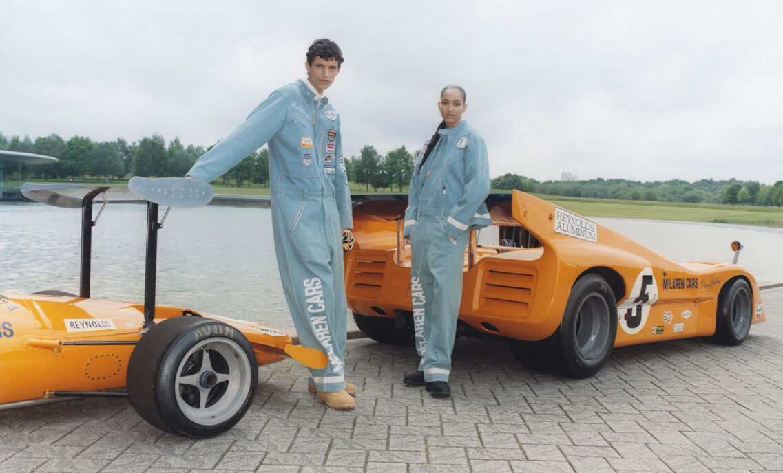 Two people pose in the Levi's® x McLaren Racing racing suits and each lean on a bright orange sports car.