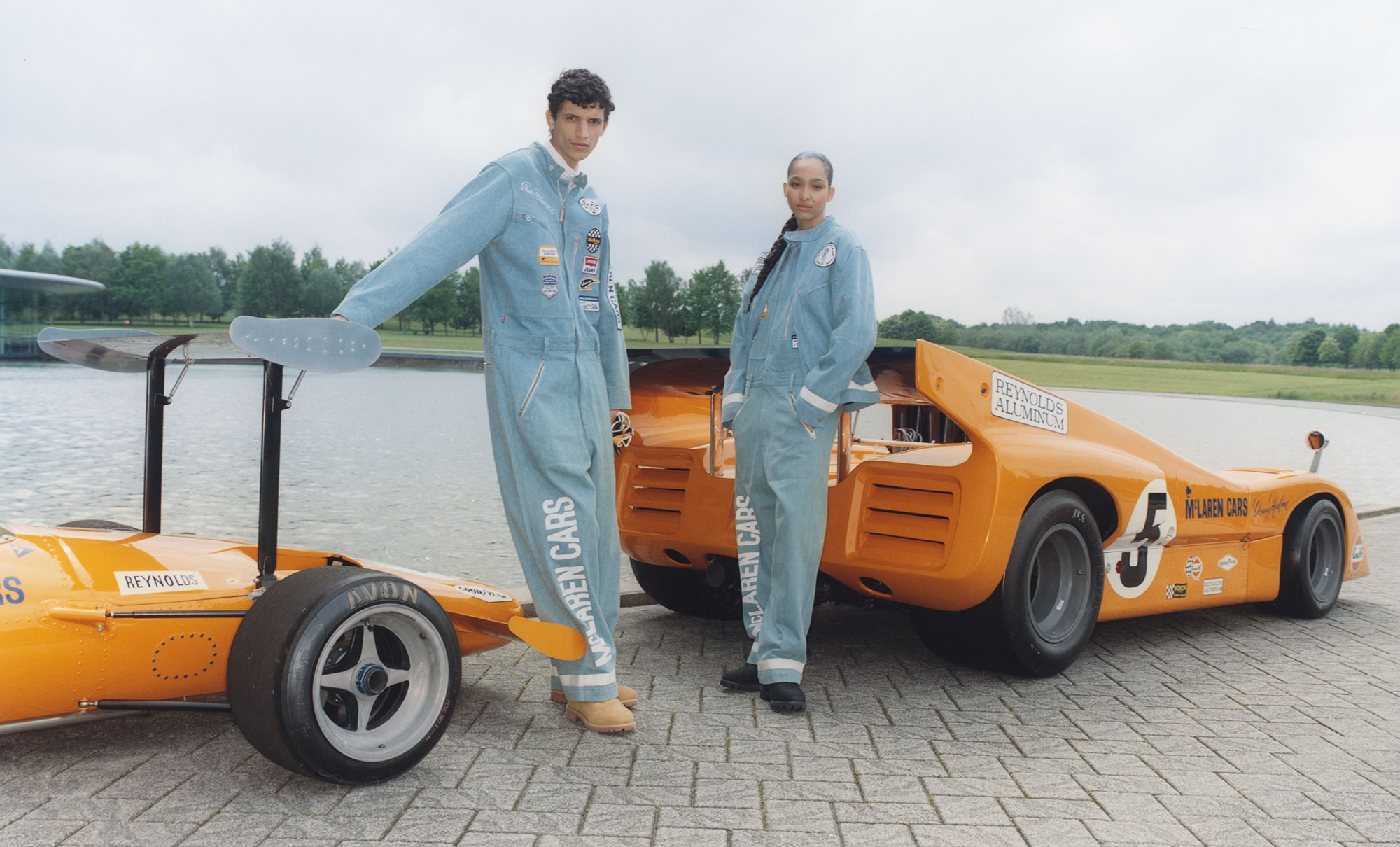 Two people pose in the Levi's® x McLaren Racing racing suits and each lean on a bright orange sports car.