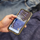 A hand holds an iPhone over two pairs of laid out jeans. The Levi's® app is opened on the phone screen, featuring a shelf of stacked jeans and the text "Even More in Store, New Store Opening, learn more."
