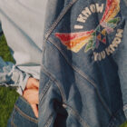 A cropped image showing the back of a person pulling on a denim jacket from the 2025 Levi's Pride collection, featuring a rainbow winged triangle and the words "I know you know"