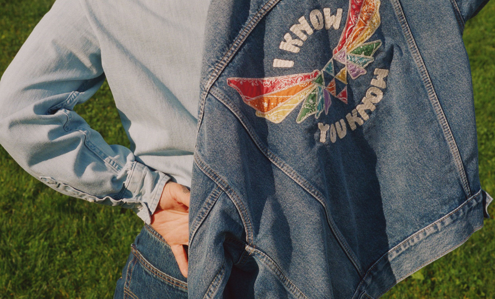 A cropped image showing the back of a person pulling on a denim jacket from the 2025 Levi's Pride collection, featuring a rainbow winged triangle and the words "I know you know"