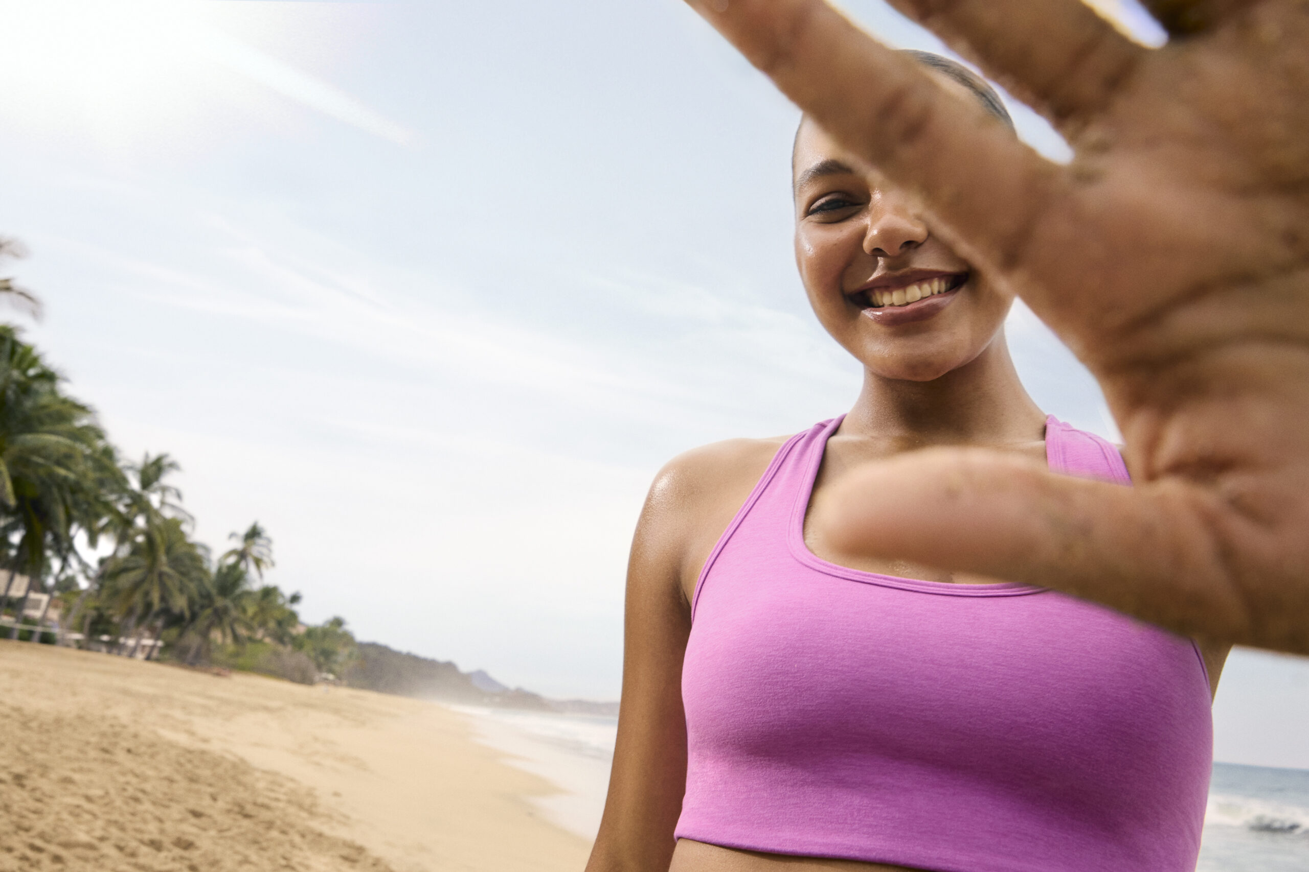 A person stands on the beach wearing a bright pink Beyond Yoga sports bra smiles with their eyes closed and their hand against the camera.