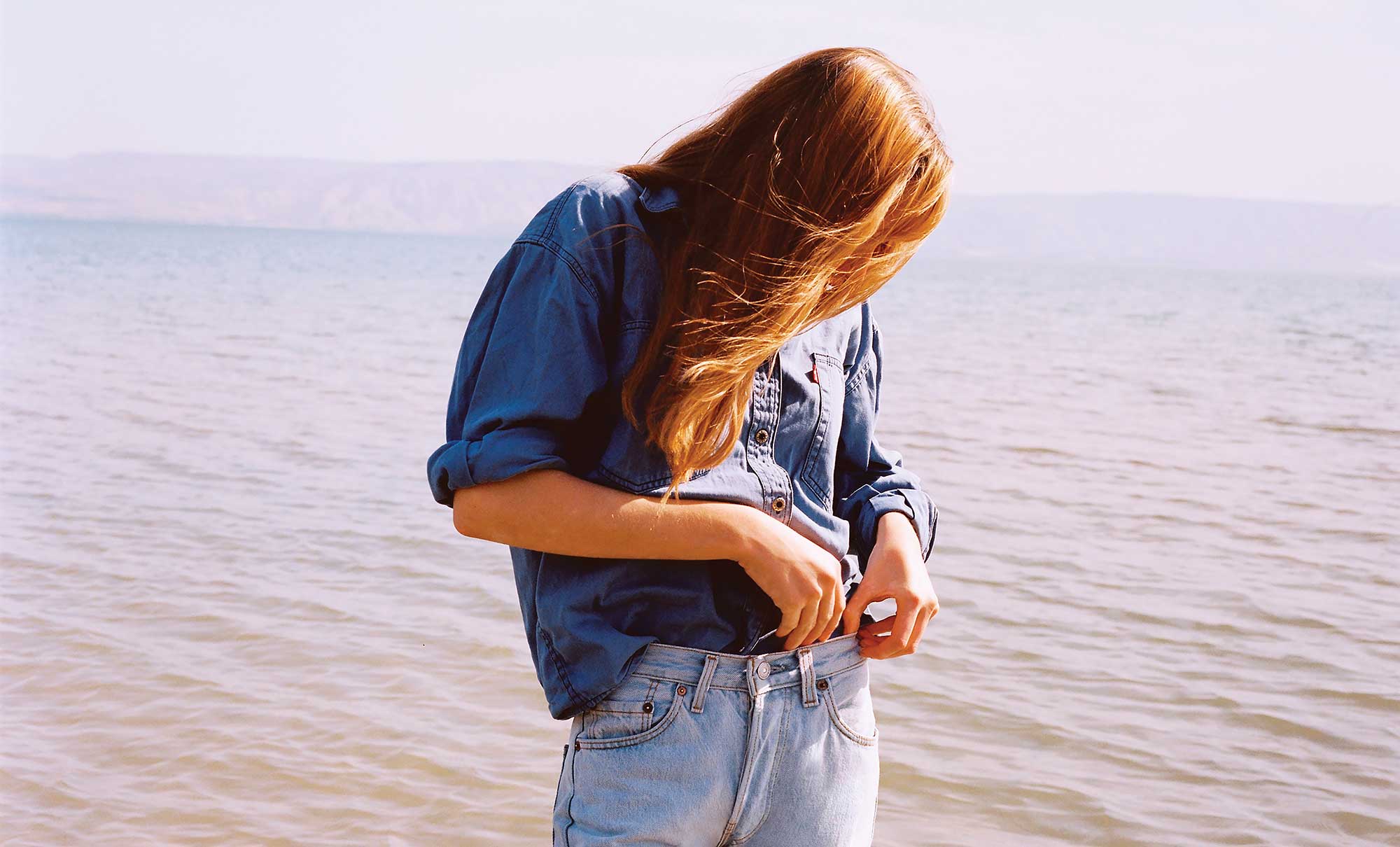 A person stands in front of an ocean with their head down and hair covering their face. They are wearing a long sleeve button down blue top with the sleeves rolled up, tucked into light wash blue denim jeans. Their hands are at their waistband.