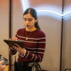 A Levi's® retail employee stands against a wall featuring a neon Levi's® batwing logo sign. They wear a red and white striped long sleeved shirt and look down at a digital tablet in their hands.