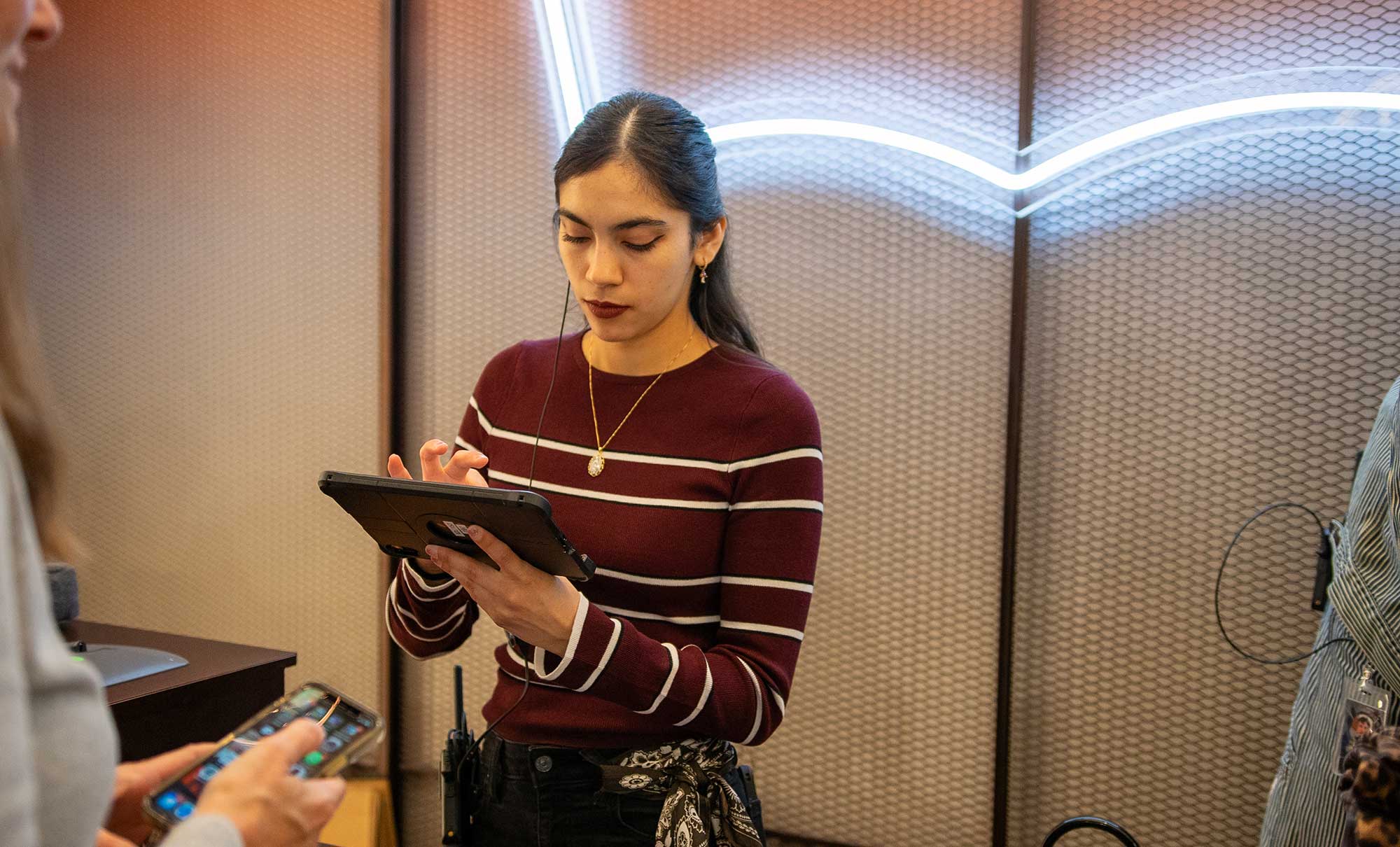 A Levi's® retail employee stands against a wall featuring a neon Levi's® batwing logo sign. They wear a red and white striped long sleeved shirt and look down at a digital tablet in their hands.