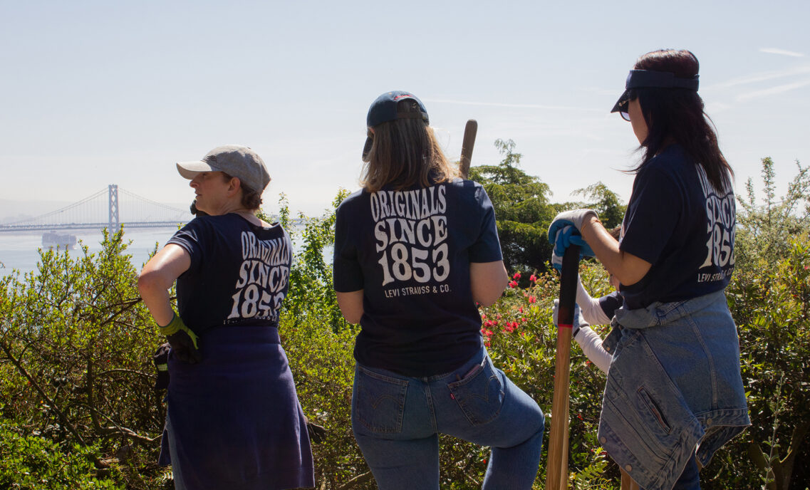Three Levi Strauss & Co. employee volunteers stand with their backs to the camera facing an overlook of the San Francisco Bay Bridge. They hold gardening tools and wear baseball caps, Levi's® jeans and blue shirts that read "Originals Since 1853 Levi Strauss & Co."